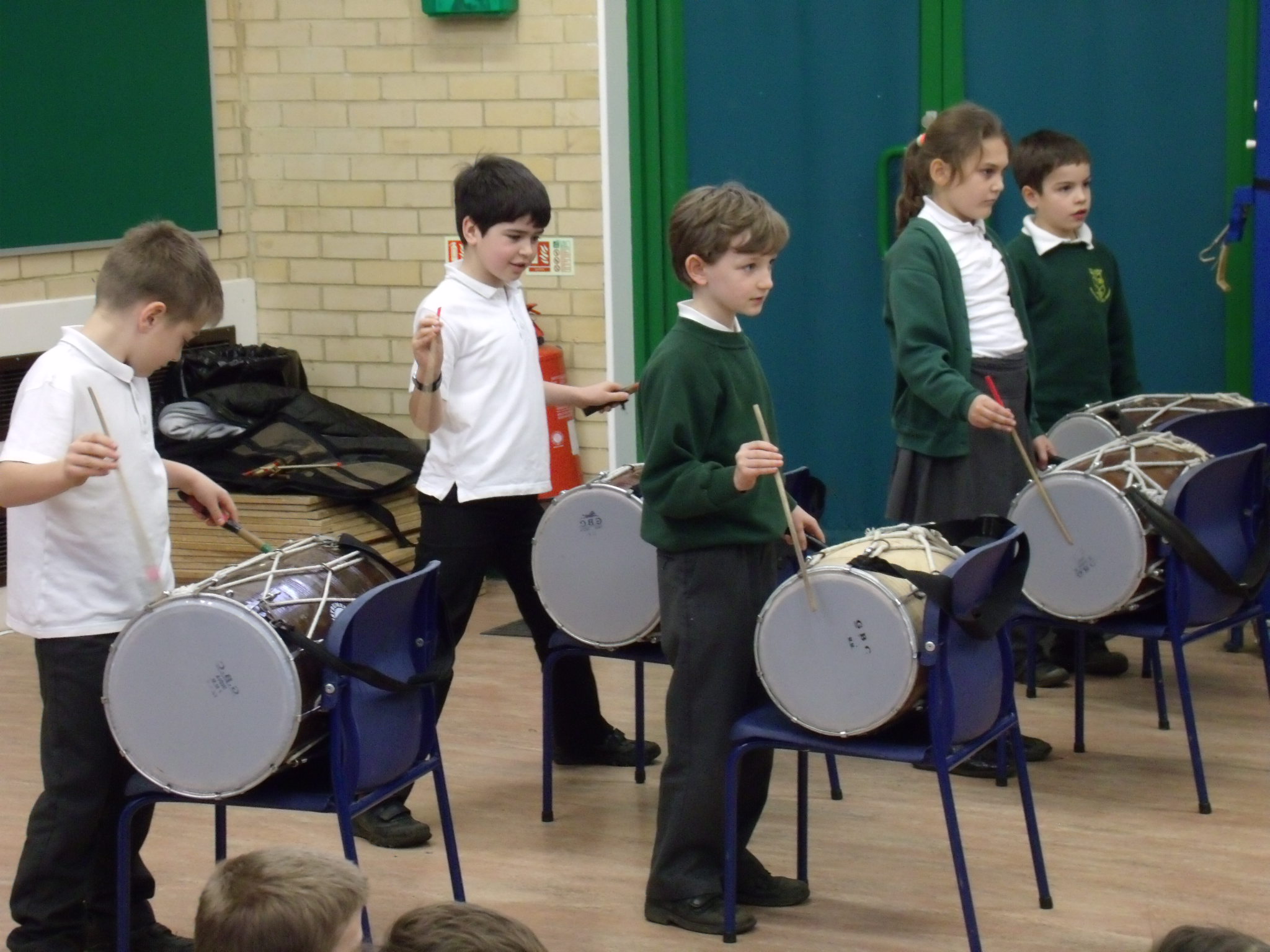 School children playing Indian drums in school hall on chairs